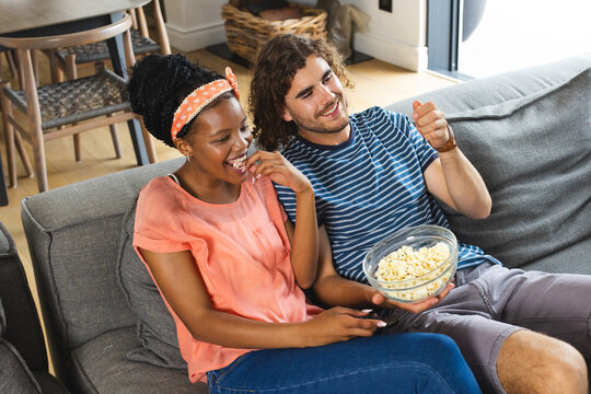 Diverse couple laughing, holding bowl of popcorn, watching television or movie at home