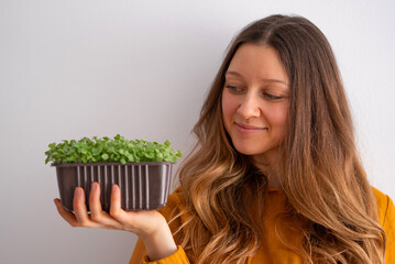 A contented young woman in a mustard yellow top admires a pot of lush green microgreens she's...