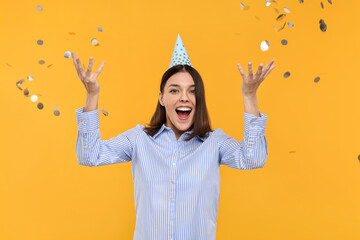 Happy young woman in party hat near flying confetti on yellow background
