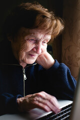 An elderly woman sits at a laptop, her face illuminated by the soft glow of the screen. With focused concentration, she types away, her wrinkled hands moving over the keyboard. - 785096106