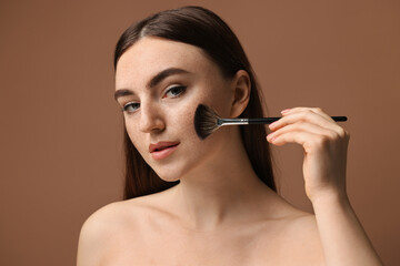 Fototapeta premium Beautiful woman with freckles applying makeup with brush on brown background