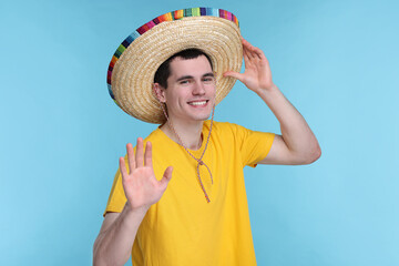 Young man in Mexican sombrero hat waving hello on light blue background