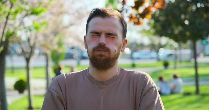 The when a middle-aged man in a park setting, through close-up shots, looks at the camera and makes gestures and facial expressions that indicate being sleepy