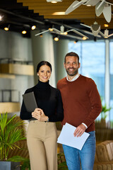 Two business colleagues posing for the camera in the office building, holding a laptop and some documents.