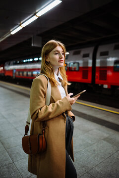 Female standing at subway platform with mobile phone, using social media and waiting train