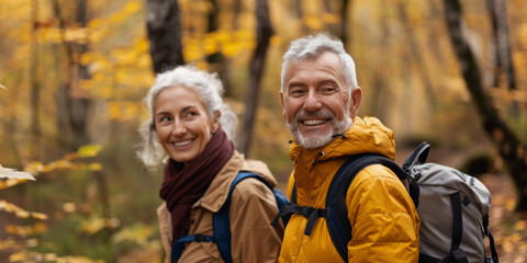 Joyful elderly couple with backpacks enjoying a hike among autumn leaves in the forest.