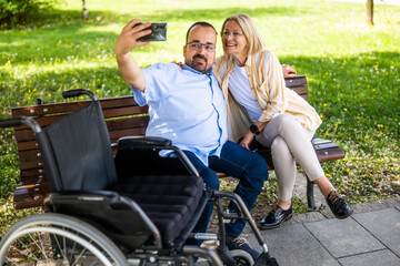 Man in wheelchair is spending time with his mother in park. They are taking selfie.