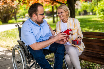 Man in wheelchair is spending time with his mother in park. He is receiving a present. - 785111387