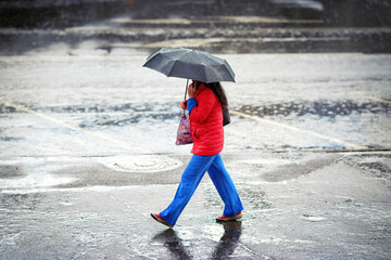 Alone woman walking with black umbrella on wet sidewalk during rain. Woman walks with umbrella through the empty streets, side view. City rain scene.