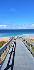 Obraz premium Vertical shot of a bridge leading to a sandy coast with the ocean in the background on a sunny day
