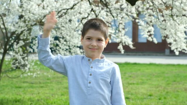 Six-year-old boy waving his hand hello or bye while standing near cherry blossom in park in spring