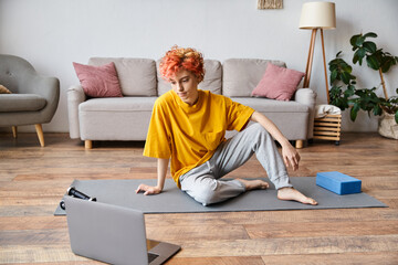 beautiful queer person in yellow t shirt sitting on yoga mat and looking at laptop before exercising