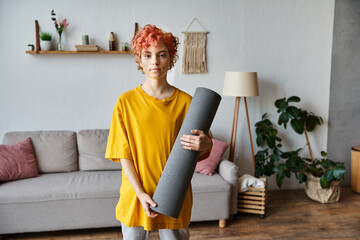 sporty young queer person with red hair in vibrant attire holding yoga mat and looking at camera