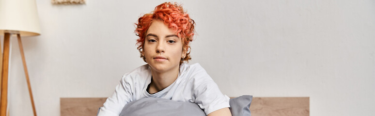 sleepy extravagant queer person in homewear with red hair sitting on bed looking at camera, banner