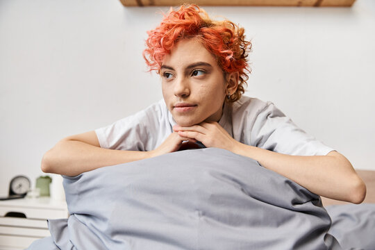 relaxing red haired queer person in homewear sitting on her bed at home and looking away, leisure