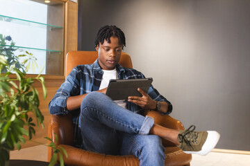 African American man sitting in chair, looking at tablet in a modern business office