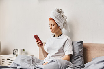 young extravagant person in homewear with hair towel looking at her smartphone while relaxing in bed