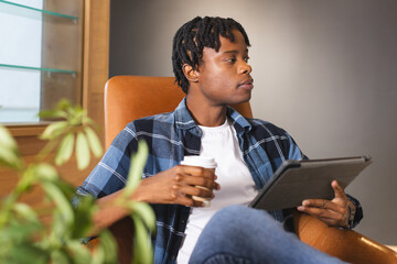 African American man holding coffee, looking at tablet in a modern business office