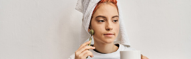 appealing queer person with hair towel using face roller and holding tea cup while at home, banner