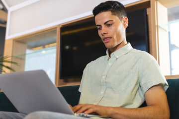 Biracial young man working on laptop, sitting on couch in a modern business office