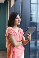 A young Asian woman holding a tablet and looking away in a modern business office