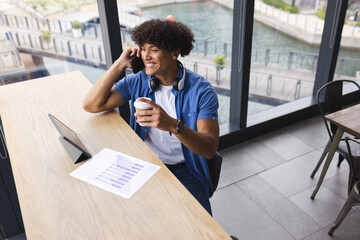 A young biracial man holding coffee, talking on the phone, sitting by a laptop in a modern business 