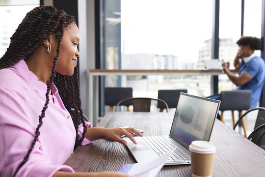 A young biracial woman with braided hair is working on a laptop in a modern business office