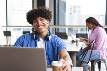 Biracial young man with headphones working on laptop in a modern business office, woman behind