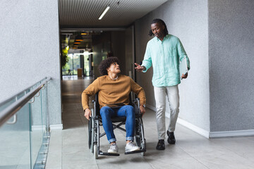 African American man pushing wheelchair of biracial man, both talking in a modern business office