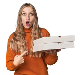 Blonde woman holding pizza boxes in studio pointing to the side