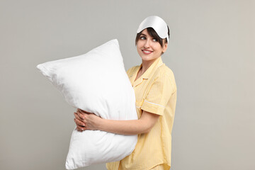 Happy woman in pyjama and sleep mask holding pillow on grey background