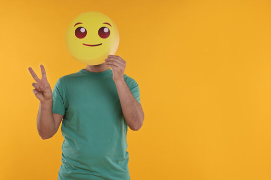 Man covering face with smiling emoticon and showing peace sign on yellow background. Space for text