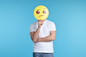 Man covering face with thinking emoticon on light blue background