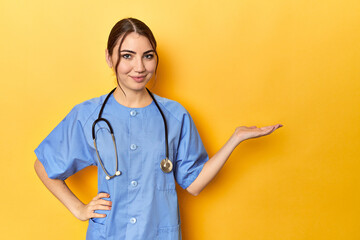 Young nurse in a yellow studio showing a copy space on a palm and holding another hand on waist.