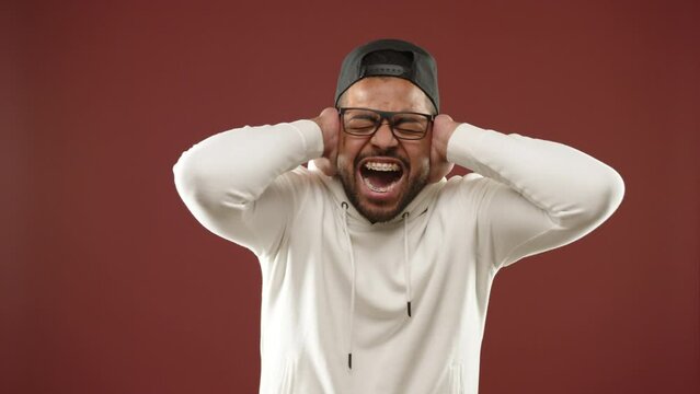 In distress, a young man with a cap and braces covers his ears with his hands, visibly pained by a loud, high decibel noise against a warm brown backdrop. High-quality 8K resolution footage. 