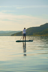 A man is standing and paddling on a paddleboard on the lake at sunset. Enjoying the vacation.  Landscape in the background. Training SUP board.