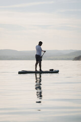 Silhouette of a man paddling on a stand up paddle boarder or SUP on the lake. Holding a paddle....