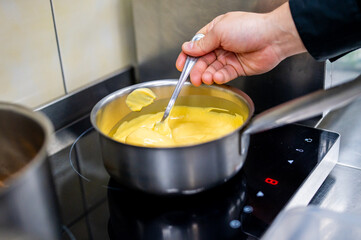 Close-up of a person’s hand stirring a creamy yellow mixture in a stainless steel pot on an induction cooktop, in a kitchen with tiled walls