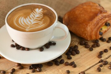 Closeup shot of a cappuccino and a freshly baked chocolatine surrounded by coffee beans