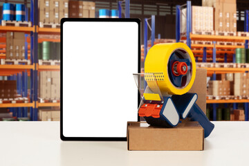 Packaging center. Scotch tape with cardboard box. Tablet computer with white screen. Dispenser with adhesive tape for packaging goods. Mock up tablet inside warehouse. Gadget near parcels for storage