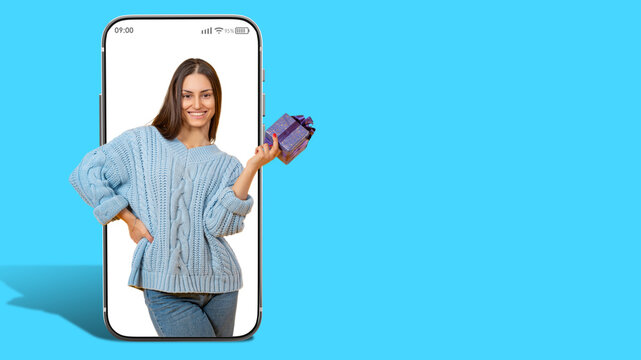 Woman with gift box in hands. Girl in screen of large phone. Promoter announces prize draw. Gift for woman birthday. Lady with gift on blue background. Concept of draw in mobile application