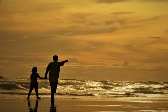 A silhouette of a father and his daughter on the beach against a backdrop of a vibrant orange sky.