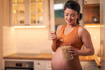 A smiling mid adult pregnant woman preparing herself a breakfast in the kitchen and holding a jar...
