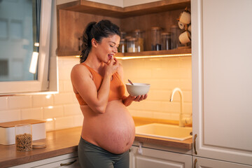 A cheerful pregnant woman having a bowl of oatmeal for breakfast in a kitchen