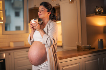 A contented pregnant woman smelling a cup of coffee and enjoying the morning in a kitchen - obrazy, fototapety, plakaty