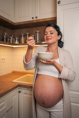 A satisfied pregnant woman enjoying her oatmeal breakfast while standing in the kitchen