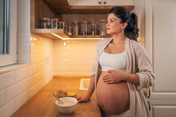 A beautiful adult pregnant woman leaning on a kitchen counter while looking outside the window