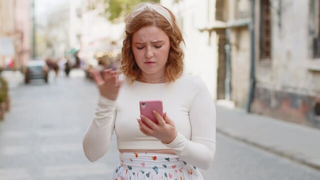 Redhead young woman in long dress use smartphone typing browsing loses becoming surprised sudden lottery results bad news fortune loss app fail virus thief fraud outdoors. Girl in urban city street.
