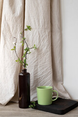 Cozy home interior decor in natural earthy colors, green clay cup with drink, ceramic vase with tree branch, wooden board on table, neutral beige linen curtain, aesthetic calm still life