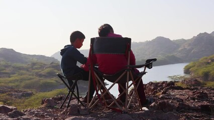 son and mother taking with each other and sitting at camping chair at mountain top with lake view